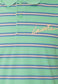 Polo vert avec des rayures horizontales bleues et beiges, deux boutons verts et la signature "Lacoste" couleur pêche sur la poitrine.