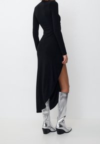 Nainen, jolla on päällään pitkä musta mekko, jossa on korkea sivureikä, ja metallinhohtoiset hopeiset polvenyli saappaat, seisoo yksinkertaista valkoista taustaa vasten.