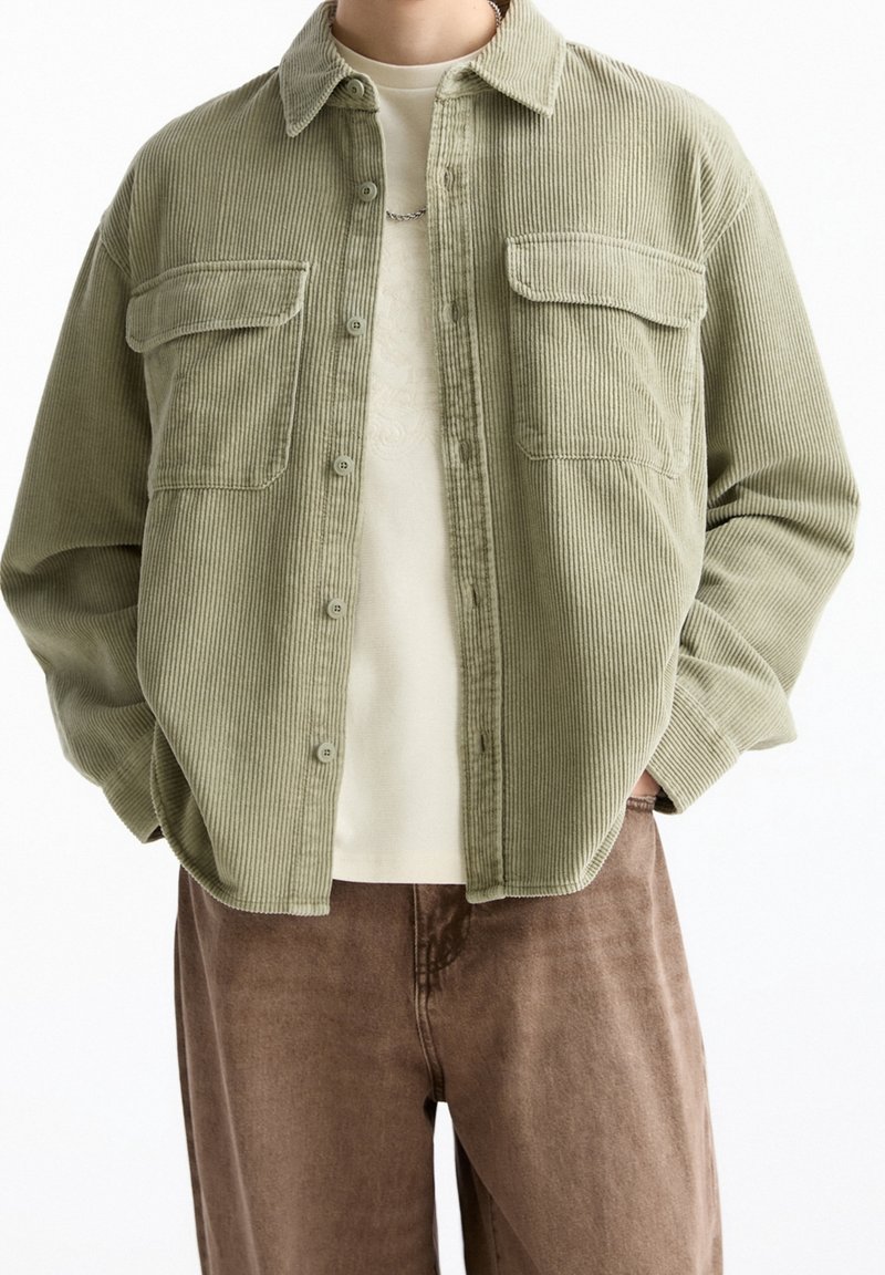 Person, die ein hellolivgrünes Cordhemd mit Knopfleiste und zwei Brusttaschen über einem beigen Shirt und braunen Hosen trägt, mit den Händen in den Taschen.