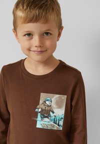 Jongentje met kort blond haar dat een bruin shirt draagt met daarop een cartoonpinguïn die snowboardt in besneeuwde bergen.