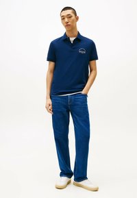 Marineblauwe polo met een klein wit logo op de borst, gecombineerd met blauwe rechte jeans en witte sneakers. Eenvoudige en casual look.
