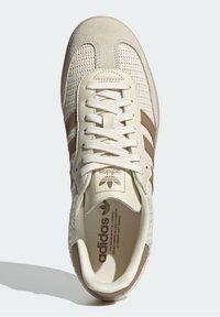 Baskets Adidas beiges avec tige en tissu tissé, trois bandes marron sur les côtés, lacets crème, et logo Adidas sur la languette et la semelle intérieure.