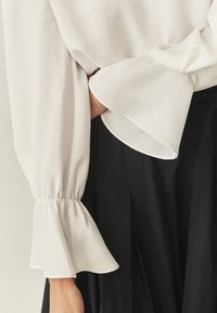 Biała, zwiewna bluzka z rozszerzanymi rękawami, wykonana z miękkiego materiału. Bluzka jest sparowana z czarną, plisowaną spódnicą z ściągaczem.