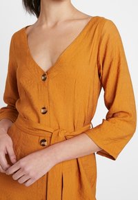 Orange, texturerad klänning med djup V-ringning, trekvartslånga ärmar, knappar ner på framsidan och en knytbälte i midjan.