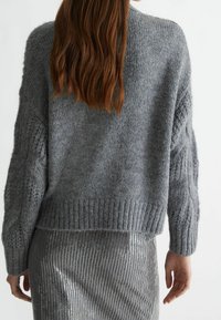 Grauer gestrickter Pullover mit überschnittenen Schultern und strukturierten Ärmeln, kombiniert mit einem figurbetonten Paillettenrock, der das Licht reflektiert.