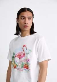 Hvid bomulds T-shirt med et farverigt flamingo-tryk omgivet af tropiske planter og blomster. Korte ærmer og klassisk pasform.
