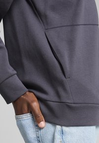 Sudadera gris oscura con un acabado texturizado y un bolsillo canguro, combinada con jeans de mezclilla azul claro. Primer plano del detalle de la manga y el bolsillo.