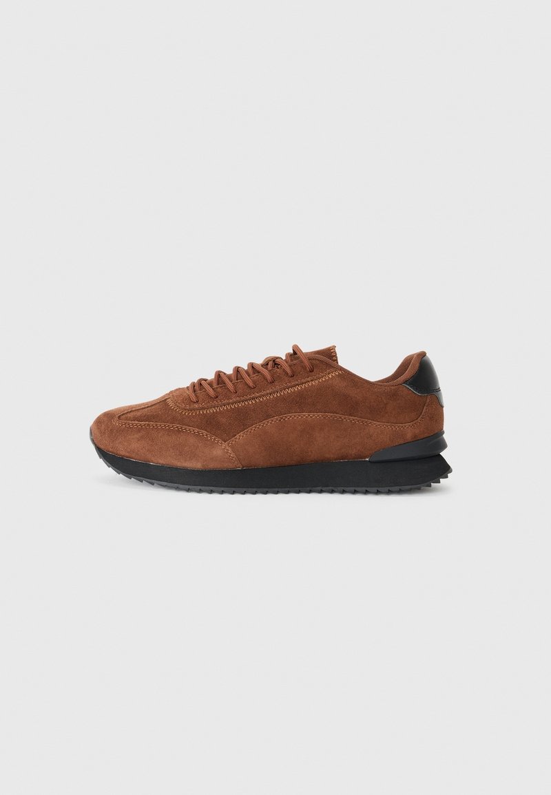 Baskets en daim marron avec semelle noire et détail au talon, fermeture à lacets et accents cousus, vue de côté sur fond uni.