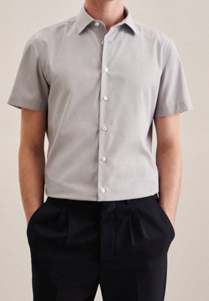 Homme portant une chemise boutonnée à manches courtes gris clair rentrée dans un pantalon noir plissé, mains dans les poches, fond neutre.