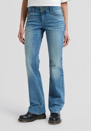 Person trägt hellblaue Bootcut-Jeans mit Doppelknopfverschluss in der Taille und schwarze Lederschuhe und steht vor einem schlichten Hintergrund.