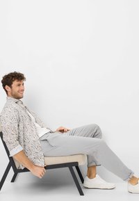 Camicia floreale leggera con motivi blu, gialli e verdi, abbinata a pantaloni grigi e sneakers bianche; seduto su una sedia moderna nera.