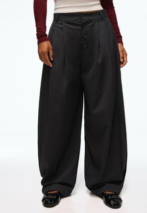 Femme portant un pantalon large noir à rayures fines avec des boutons à l'avant, des chaussures plates noires et un haut à manches longues bordeaux et blanc.