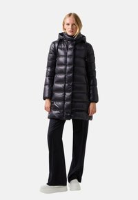 Doudoune en tissu noir brillant, dotée d'une capuche, d'une fermeture éclair et de panneaux matelassés horizontaux. Associée à un pantalon foncé et des chaussures blanches.