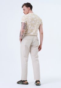 Chemise beige à manches courtes avec motif de feuilles de palmier, associée à un pantalon beige clair et des sandales multicolores, vue de dos.