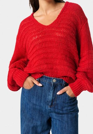 Maglione rosso lavorato a maglia con scollatura a V, texture a costine e maniche over size, abbinato a jeans in denim blu a vita alta con tasche anteriori.