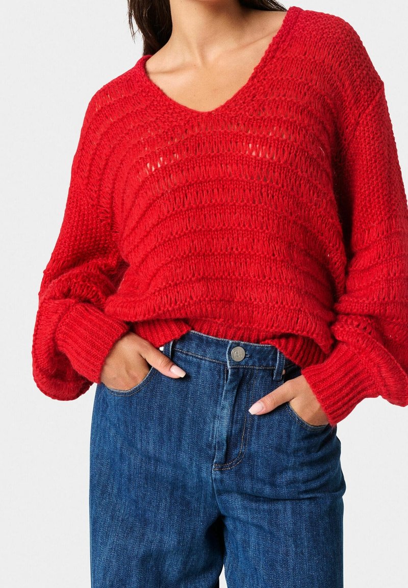 Czerwony sweter z dzianiny, z dekoltem w serek, prążkowaną fakturą i oversize'owymi rękawami, w zestawieniu z niebieskimi dżinsami z wysokim stanem, posiadającymi kieszenie z przodu.