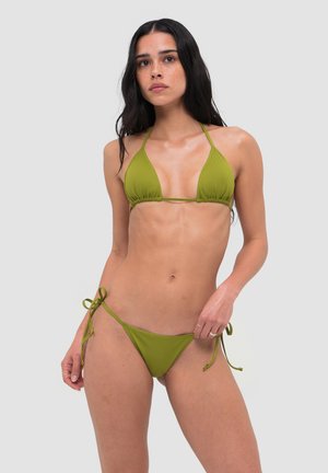 Kobieta z długimi ciemnymi włosami, ubrana w oliwkowo-zielony bikini na sznureczki z wiązaniami po bokach na jednolitym jasnym tle.