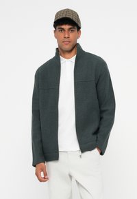 Casaco verde escuro com fecho de correr, feito de tecido texturizado, apresentando um colarinho alto e duas costuras horizontais no peito. Combine com uma polo branca e calças claras.