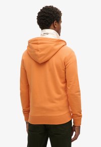 Mand vender væk iført en orange hoodie over en hvid skjorte med en synlig "Superdry"-logo på kraven.