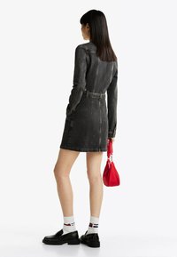 Abito di jeans scuro con maniche lunghe, vita sottile e silhouette aderente. Il modello tiene una piccola borsa rossa, abbinata a scarpe nere e calze bianche.