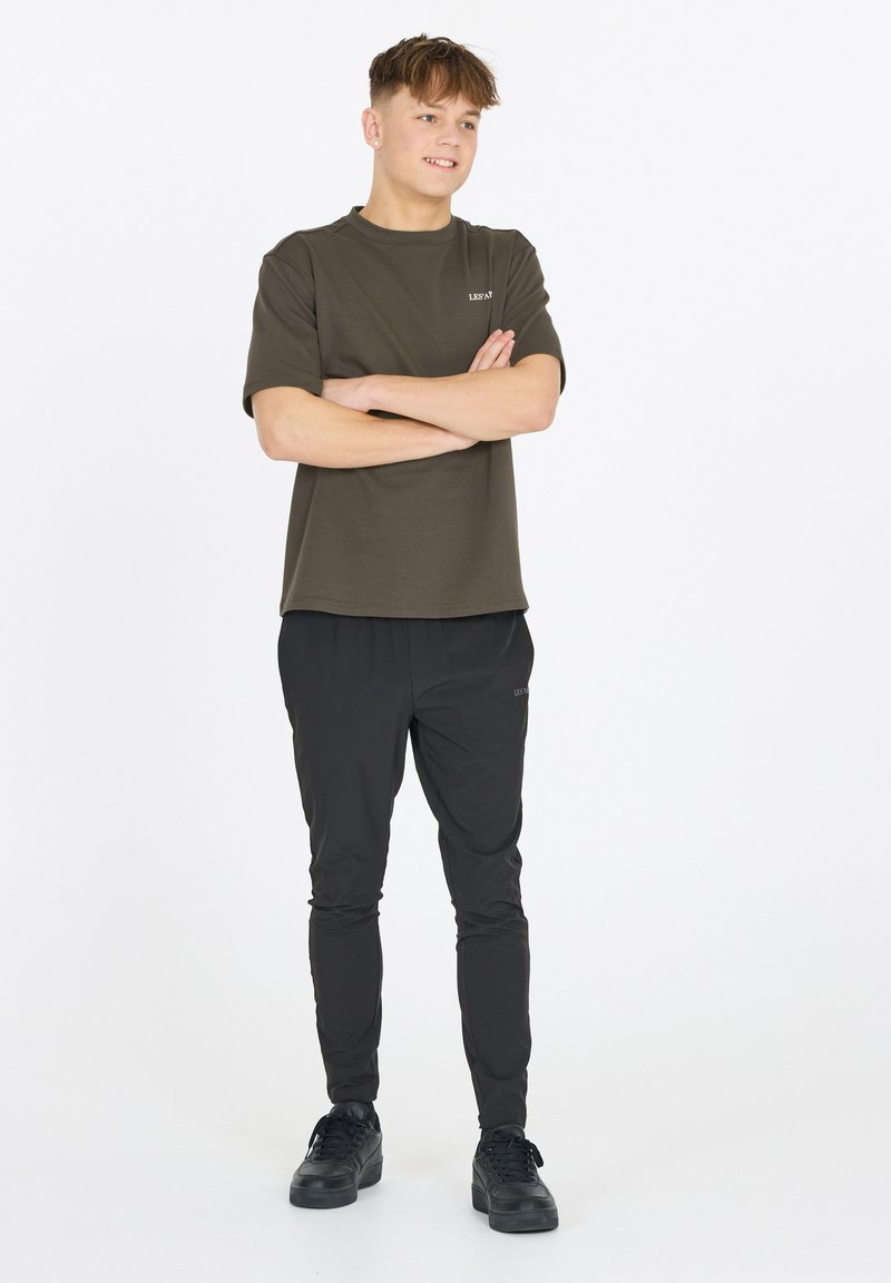 Oliivinvihreä t-paita lyhyillä hihoilla ja pyöreällä kauluksella, yhdistettynä mustiin urheiluhousuihin. Mustat lenkkarit täydentävät asun.