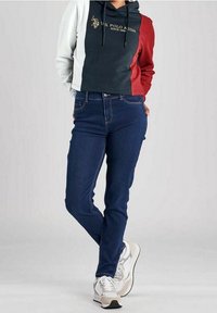 Person som bär en tri-färgad U.S. Polo Assn. hoodie, mörkblå jeans och vita sneakers, stående med benen korsade.