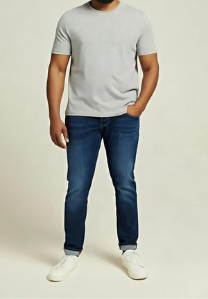Homme portant un t-shirt gris clair à manches courtes, un jean bleu foncé retroussé et des baskets blanches, debout avec une main dans la poche.