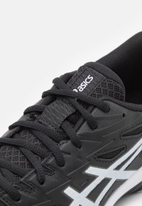 Chaussure de sport noire avec un dessus en mesh, des superpositions texturées, des lacets plats et un logo blanc sur le côté. Design élégant avec des détails prononcés.