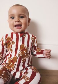 Czerwono-biały pajacyk w paski z wzorem pierniczków. Miękka tkanina, długie rękawy, zamek błyskawiczny z przodu oraz dopasowany krój.