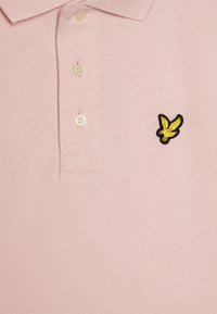 Pikéskjorte i lys rosa, med teksturert stoff, to knapper, og en liten, svart og gul brodert ørnerlogo på brystet.