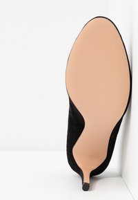 Chaussure noire à talon haut reposant contre un mur blanc, montrant une semelle beige lisse avec un bout pointu et un talon fin.