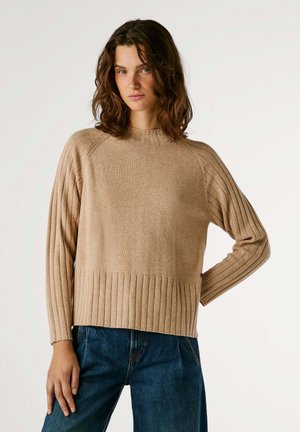 Beige gebreide trui met een geribbelde textuur, ronde hals en lange mouwen. Kenmerkt zich door een zoom met verticale ribbels, gecombineerd met blauwe jeans.