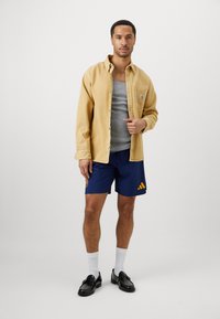 Camisa mostarda de botões sobre uma t-shirt cinza, calções azul-marinho com detalhes amarelos, sapatos pretos e meias brancas.