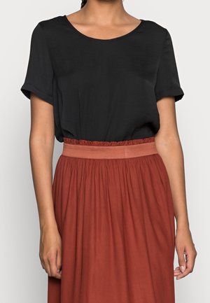 Kvinde iført en kortærmet sort top, stoppet ned i et højtaljet, rustfarvet sammensyet skørt med en tekstureret talje.