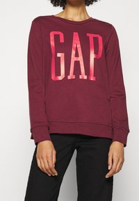 Sudadera burdeos con un gran logotipo "GAP" en rosa. Fabricada con una tela suave, tiene un corte relajado y puños acanalados, con un cuello redondo.