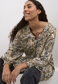 Smiješna žena s dugom kovrčavom kosom koja nosi bež i crnu bluzu s uzorkom cvijeća i tamne hlače, sjedi naslonjena na svijetlo sivu pozadinu.