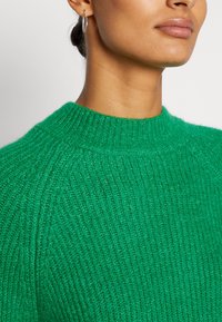 Zielony sweter z dzianiny z wysoką szyją, prążkowaną teksturą oraz dopasowanym krojem. Zbliżenie ukazujące materiał i kolor.