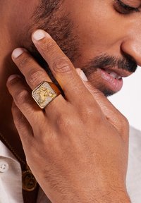 Bague hexagonale en or avec une surface texturée mettant en avant un design d'aigle en relief, portée sur une main avec des ongles courts et bien entretenus.