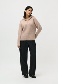 Kvinne med svart hår har på seg en beige strikkegenser med glidelås foran, svarte vide bukser og svarte sko, stående mot en ensfarget bakgrunn.