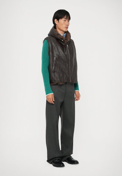Man wearing dark brown leather vest with fur lining, teal long-sleeve shirt, gray wide-leg trousers, and black shoes, standing against white background.