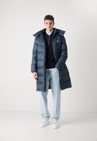 Marineblauer Puffermantel mit Kapuze, ausgestattet mit einem Reißverschluss, Seitentaschen und einem Logo-Akzent, kombiniert mit hellblauen Jeans und weißen Turnschuhen.
