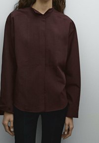 Chemise bourgogne à manches longues avec col mandarin, doté de plis verticaux sur le devant, fabriquée en tissu doux. Fermeture à boutons au niveau du cou.