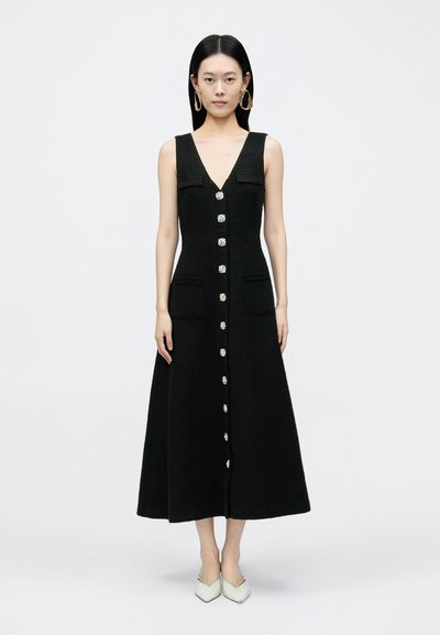 Schwarzes ärmelloses Kleid mit V-Ausschnitt, vorderer Knopfleiste, zwei Brusttaschen und ausgestellter Silhouette. Das Model trägt hellfarbene High Heels.