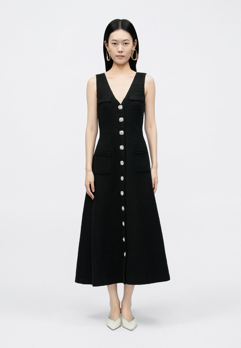 Melna piedurkņu kleita ar V veida izgriezumu, priekšpusē ar pogām, divām krūšu kabatām un paplašinātu siluetu. Modelis valkā gaismas krāsas kurpes ar papēžiem.