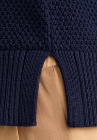 Granatowy dzianinowy tekstyl z fakturowanym wzorem, z prążkowanym obszyciem na dole. U dołu widoczny otwór w kształcie klucza. Pod spodem beżowa tkanina.