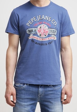 Mand iført en blå Pepe Jeans Co. T-shirt med et Union Jack-emblem og falmede blå jeans.