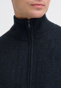 Schwarzer Pullover mit hohem Kragen und Reißverschluss, der ein strukturiertes Strickmuster und einen gerippten Kragen zeigt. Der Stoff wirkt weich und warm.