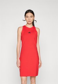 Raudona be rankovių suknelė, pagaminta iš lygios medžiagos, su prigludusia konstrukcija, kurią puošia mažas juodas Adidas logotipas ant krūtinės.