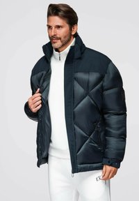 Marinblå pufferjacka med diamantquiltat mönster, hög krage, dragkedja och sidofickor, bärs över en vit tröja med ståkrage.