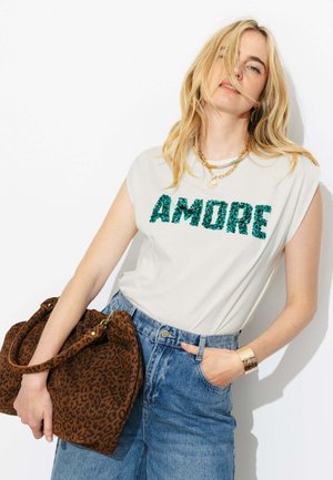 Femme aux longs cheveux blonds porte un top blanc sans manches avec "AMORE" en sequins verts, un jean bleu, des bijoux en or et tient un sac marron imprimé léopard.